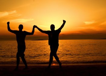 Dos hombres bailando a las orillas del mar bajo la puesta del sol. Fuente: Dana Tenis / Pexels.com.