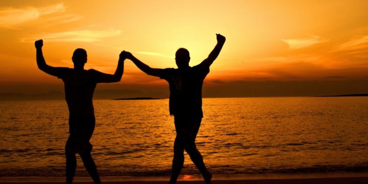 Dos hombres bailando a las orillas del mar bajo la puesta del sol. Fuente: Dana Tenis / Pexels.com.