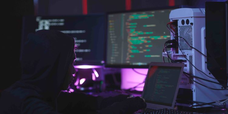 Portrait of unrecognizable hacker using computer equipment with programming code on screens in dark room, cyber security concept, copy space. Fuente: