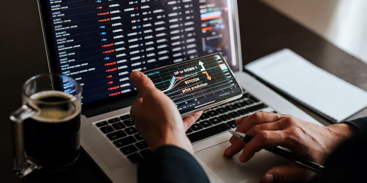 Hombre evaluando el mercado de las criptomonedas en la pantalla de un teléfono y en la computadora. Fuente: avanti_photo /   elements.envato.com