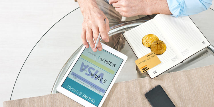 Hombre revisa cuenta bancaria con tarjeta y monedas de bitcoin sobre mesa con logo de Visa. Composición por CriptoNoticias. Photology75 / elements.envato.com; Visa / wikipedia.org; Pressmaster / elements.envato.com.