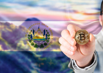 Hombre muestra moneda de Bitcoin con bandera de El Salvador sobre paisaje.