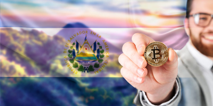 Hombre muestra moneda de Bitcoin con bandera de El Salvador sobre paisaje.