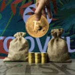 Una mano sostiene una moneda de bitcoin y debajo hay dos sacos llenos de monedas. Al fondo aparece el logo de Tropykus.