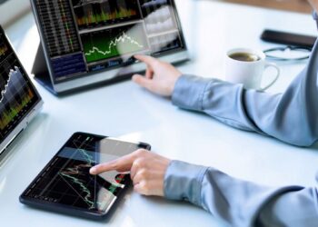 Persona sentada frente a 2 computadores y una tablet con gráficos de precios.