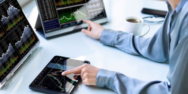 Persona sentada frente a 2 computadores y una tablet con gráficos de precios.