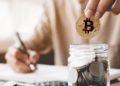 Una persona guarda una moneda de bitcoin en un vaso con múltiples monedas, mientras toma nota con un lápiz.