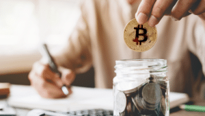 Una persona guarda una moneda de bitcoin en un vaso con múltiples monedas, mientras toma nota con un lápiz.