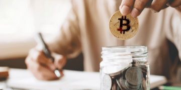 Una persona guarda una moneda de bitcoin en un vaso con múltiples monedas, mientras toma nota con un lápiz.