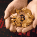 Mano sosteniendo oro y Bitcoin con gráfico rojo en el fondo.