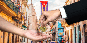 Una persona entrega una moneda física de bitcoin a otra persona y al fondo se observa una bandera de Cuba