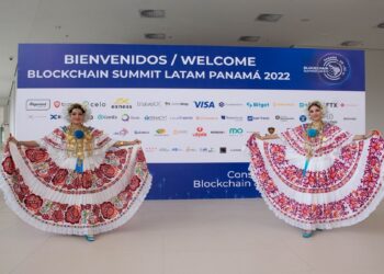 Dos mujeres con trajes típicos bailan frente a una pancarta de la Blockchain Summit LatAm 2022