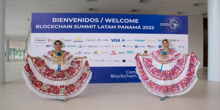 Dos mujeres con trajes típicos bailan frente a una pancarta de la Blockchain Summit LatAm 2022