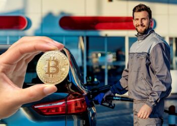 Mano muestra moneda de bitcoin con hombre cargando gasolina a su automovil en el fondo