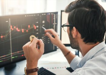 trader sosteniendo moneda de bitcoin mientras señala gráfico de precio en pantalla de computadora