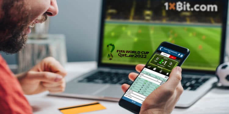 hombre siguiendo apuestas en teléfono con mundial de futbol transmitiendo en pantalla de computadora en el fondo con logo de 1xBit y mundial de futbol qatar 2022
