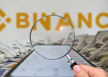 mano sosteniendo lupa sobre teléfono sobre billetes de dólares con logo de Binance en el fondo