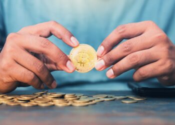 hombre sostiene entre manos moneda de bitcoin con monedas esparcidas sobre mesa