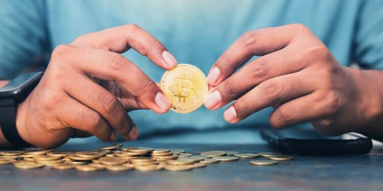 hombre sostiene entre manos moneda de bitcoin con monedas esparcidas sobre mesa