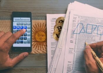 persona calculando impuestos y llenando formularios con logo de la AFIP con monedas de bitcoin escondidas entre hojas y bandera de argentina pintada sobre la mesa