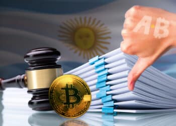 mano mostrando señal de disgusto con logo de Bitcoin Argnetina hacia moneda de bitcoin apoyada en martillo de juez junto a documentos legales con bandera de Argentina en el fondo