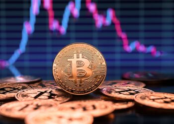 moneda de bitcoi entre otras monedas de bitcoin disperzadas sobre mesa con gráfico de precio bajista en el fondo