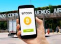 mano sosteniendo telefono movil con interfaz de bitcoin en pantalla y mercado central de buenos aires en el fondo