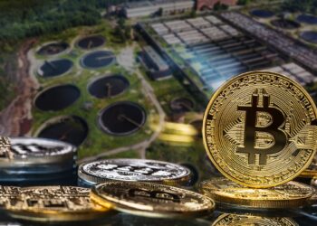 monedas de bitcoin sobre mesa con imagen aerea de planta de tratamiento de aguas residuales en el fondo