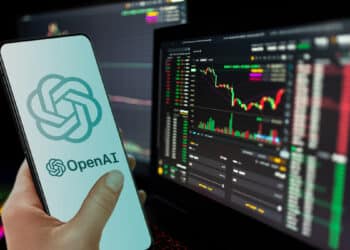 mano sosteniendo telefono movil con logo de OpenAI con pantallas de computadoras con graficos de precio