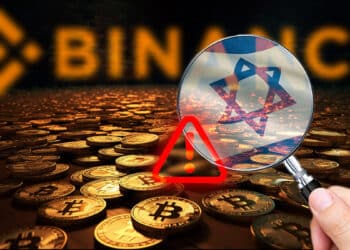 mano sosteniendo lupa con bandera de Israel sobre monedas de bitcoin esparcidas sobre habitacion oscura con logo de binance en el fondo mostrando señal de alerta