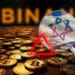 mano sosteniendo lupa con bandera de Israel sobre monedas de bitcoin esparcidas sobre habitacion oscura con logo de binance en el fondo mostrando señal de alerta