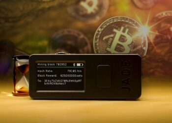 Jade Wallet sobre mesa con reloj de arena y monedas de bitcoin siendo minadas en el fondo