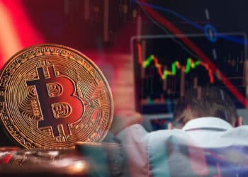 moneda de bitcoin en rojo junto a trader arrepentido frente a computadora con grafico bajista de precio en pantalla y velas de precio superpuestas.