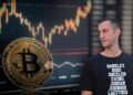 Soctt Melker observando moneda de bitcoin apoyada en pantalla con grafico de precio
