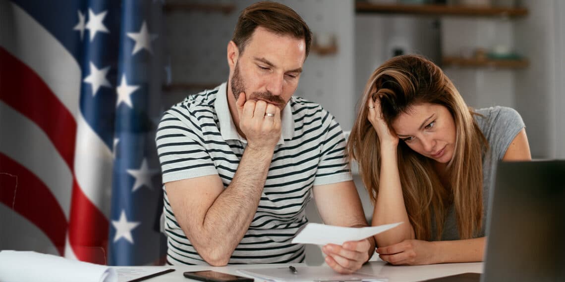 pareja preocupada observando cheque con bandera de Estados Unidos en el fondo