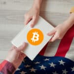 una manos sosteniendo un documento en el que se observa el logo de bitcoin y de fondo la bandera de Estados Unidos.