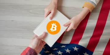 una manos sosteniendo un documento en el que se observa el logo de bitcoin y de fondo la bandera de Estados Unidos.