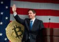Ron de Santis en campaña política con moneda de bitcoin detrás