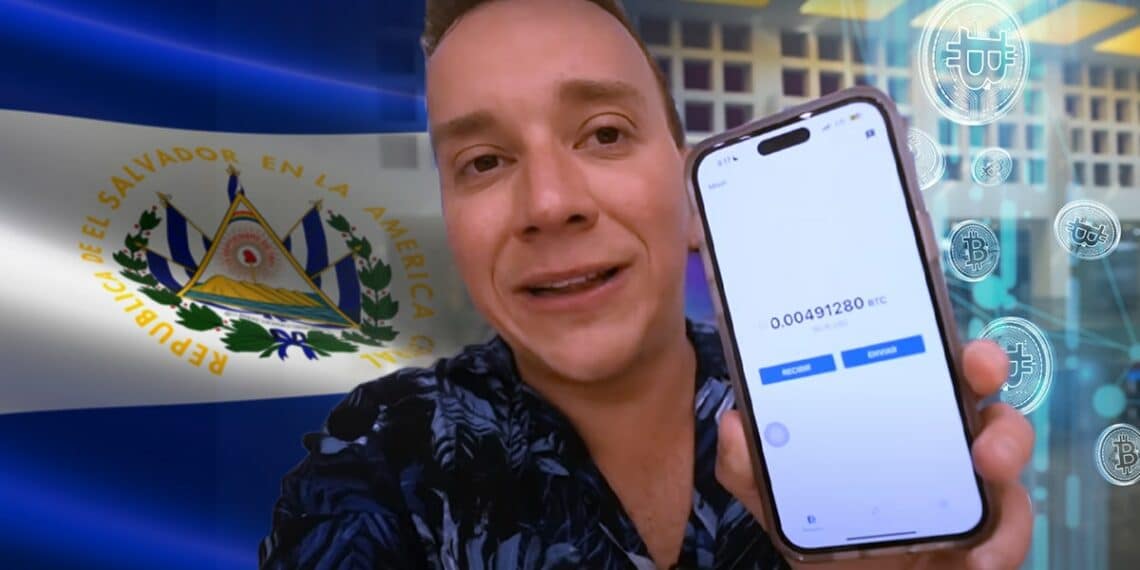 youtuber venezolano, oscar alejando sosteniendo telefono movil con billetera de bitcoin en pantalla, bandera de El Salvador y monedas digitales de bitcoin cayendo en el fondo