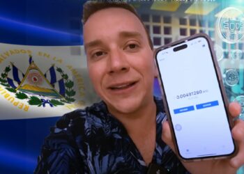 youtuber venezolano, oscar alejando sosteniendo telefono movil con billetera de bitcoin en pantalla, bandera de El Salvador y monedas digitales de bitcoin cayendo en el fondo