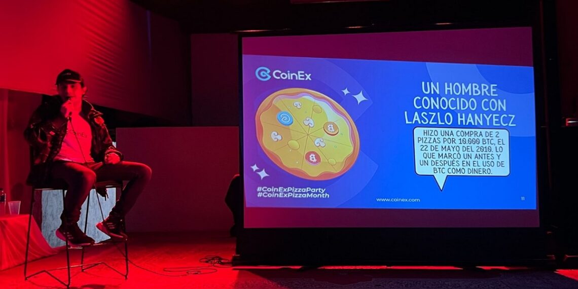 Un orador sentado en el escenario al lado de una pancarta del evento Bitcoin Pizza Party de CoinEx.