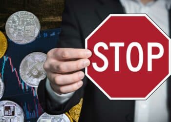 mano sosteniendo señal de alto con bitcoins y criptomonedas sobre mesa con grafico de precio en el fondo
