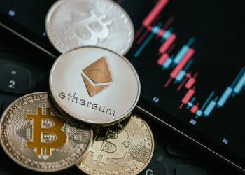 moneda de ethereum sobre monedas de bitcoin junto a pantalla con grafico de precio