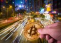 Una imagen con una moneda de bitcoin sostenida por una persona y con las vías de tránsito de una ciudad de fondo.