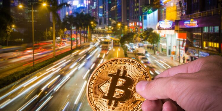 Una imagen con una moneda de bitcoin sostenida por una persona y con las vías de tránsito de una ciudad de fondo.
