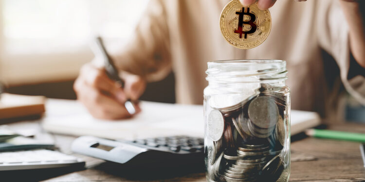 persona colocando moneda de bitcoin en frasco con monedas y haciendo cuentas en el fondo