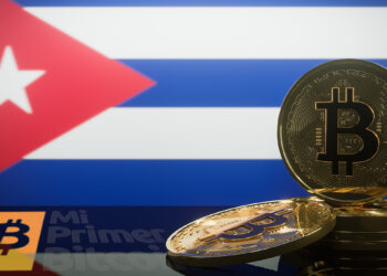 monedas de bitcoin con pantalla proyectando bandera de Cuba en el fondo y logo de Mi primer Bitcoin reflejado en mesa