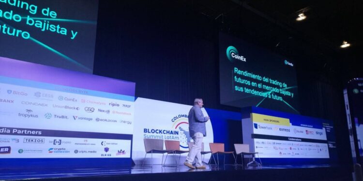 Un hombre de traje camina sobre el escenario de la Blockchain Summit Latam con imagenes de CoinEx detras.