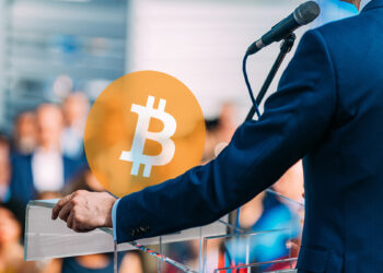 logo de Bitcoin sobre mesa con hombre dando discurso ante un publico