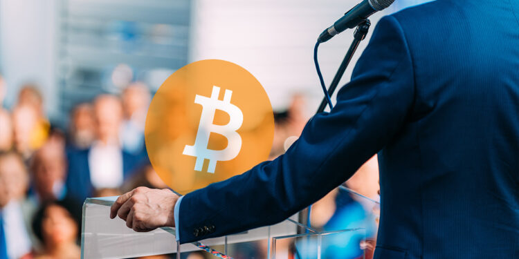 logo de Bitcoin sobre mesa con hombre dando discurso ante un publico